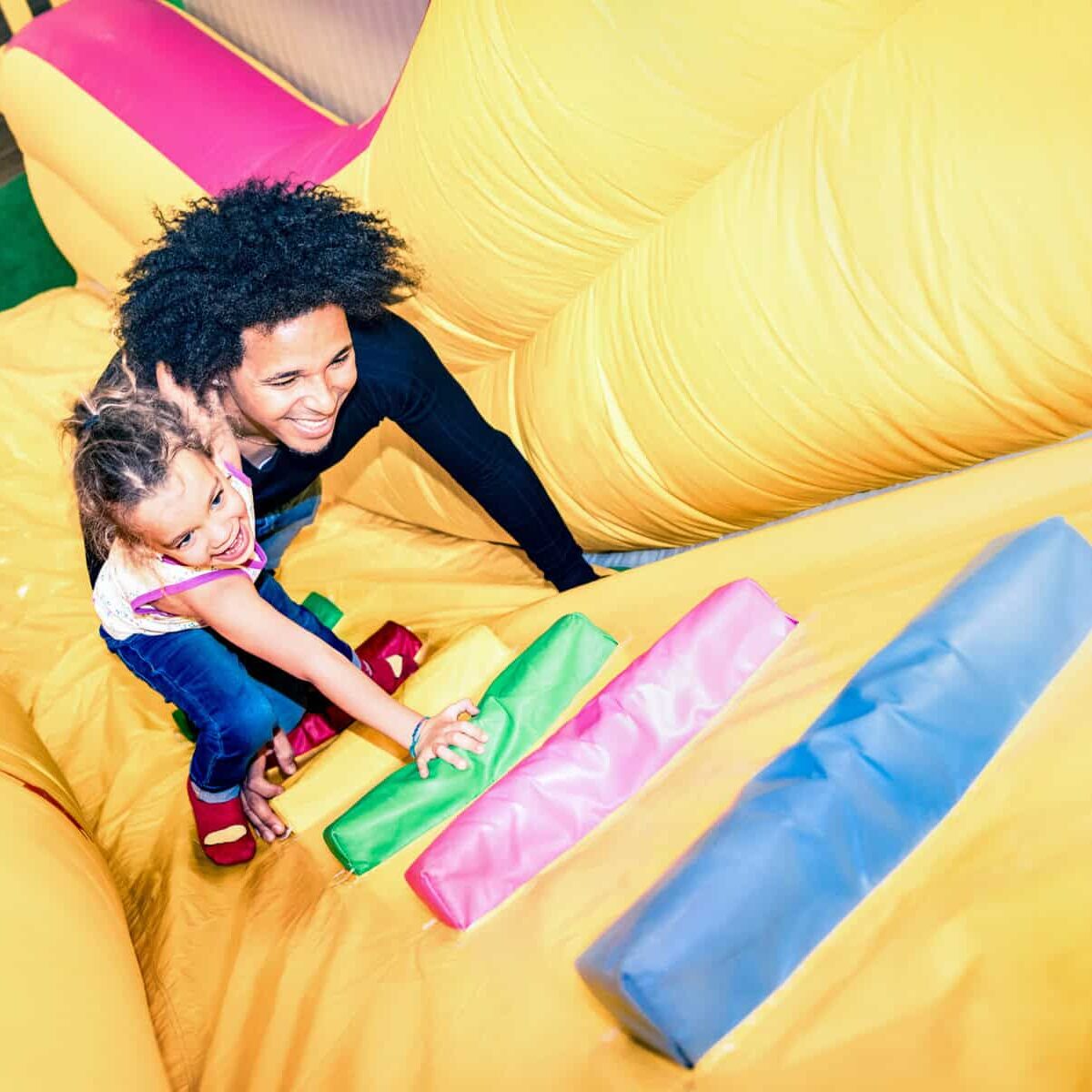 Latin american dad playing with mixed race daughter on inflatable slide at kindergarten playroom - Family concept with happy multiracial child and father having fun together at kids playground toyroom